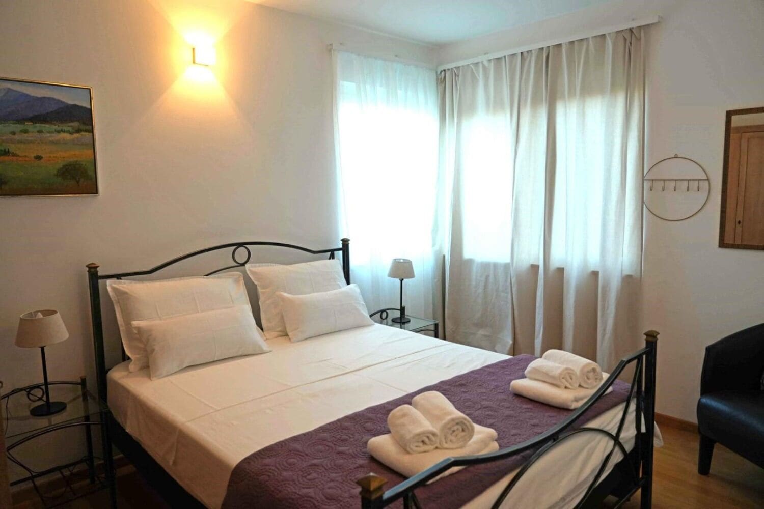 A neatly made bed with white linens, a purple blanket, and rolled towels on a metal frame bed in a room with white walls, a painting, and a window with curtains.