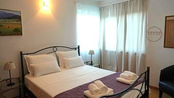 A neatly made bed with white linens, a purple blanket, and rolled towels on a metal frame bed in a room with white walls, a painting, and a window with curtains.