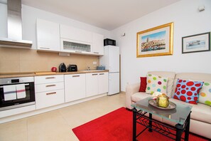 A modern kitchen with white cabinets, a Bosch oven, and a microwave. There is a red rug, a glass coffee table, and a beige sofa with colorful polka dot pillows. - Apartments Apartmani Oh La La (Supetar)