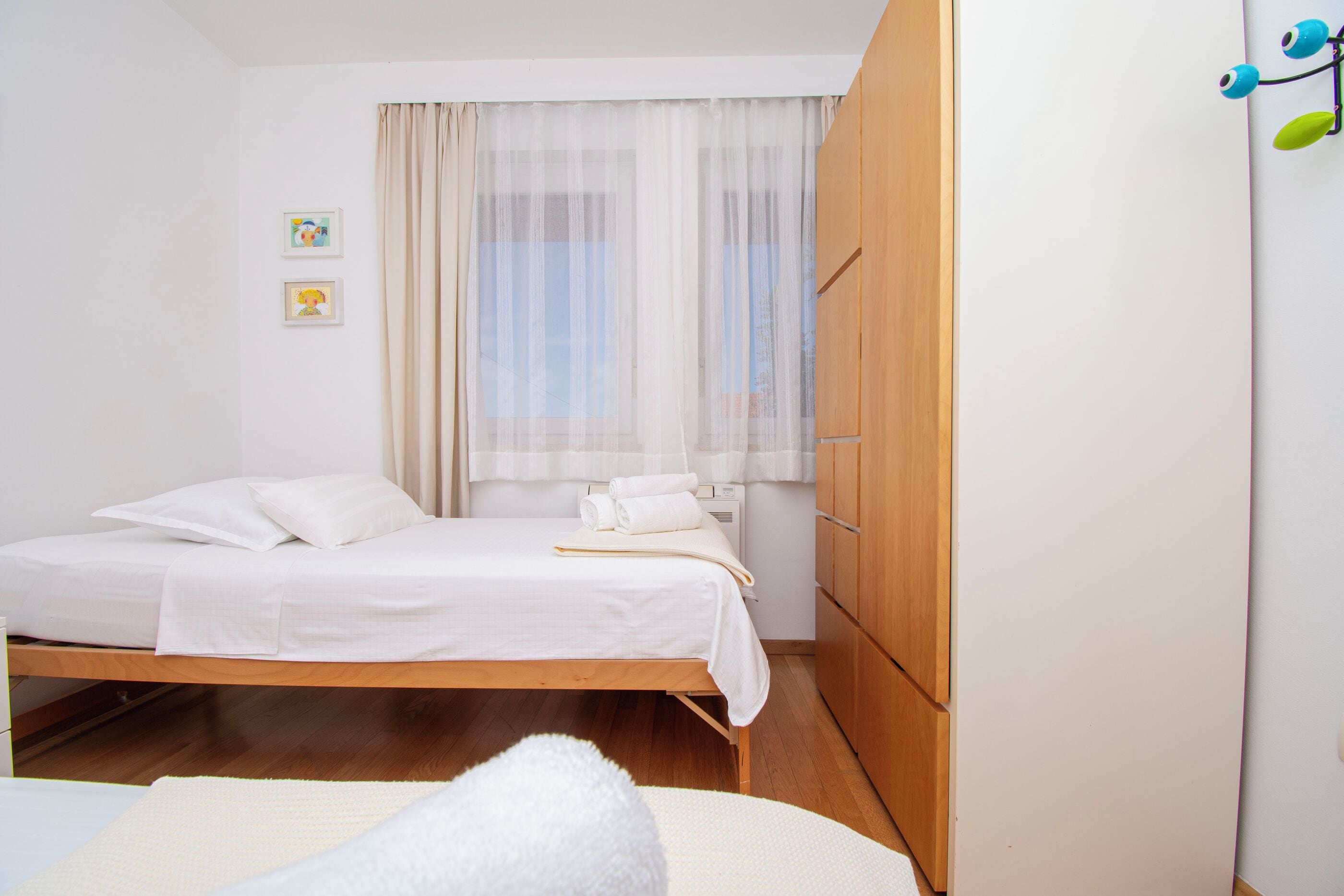 A single-bed room with a wooden wardrobe, a window with sheer curtains, and a small bedside table with a book and towels.