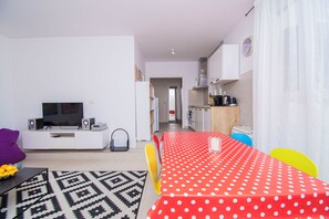 A modern living room with a TV, speakers, and a patterned rug. A dining area with a polka dot tablecloth and chairs is visible. - Apartments Apartmani Oh La La (Supetar)