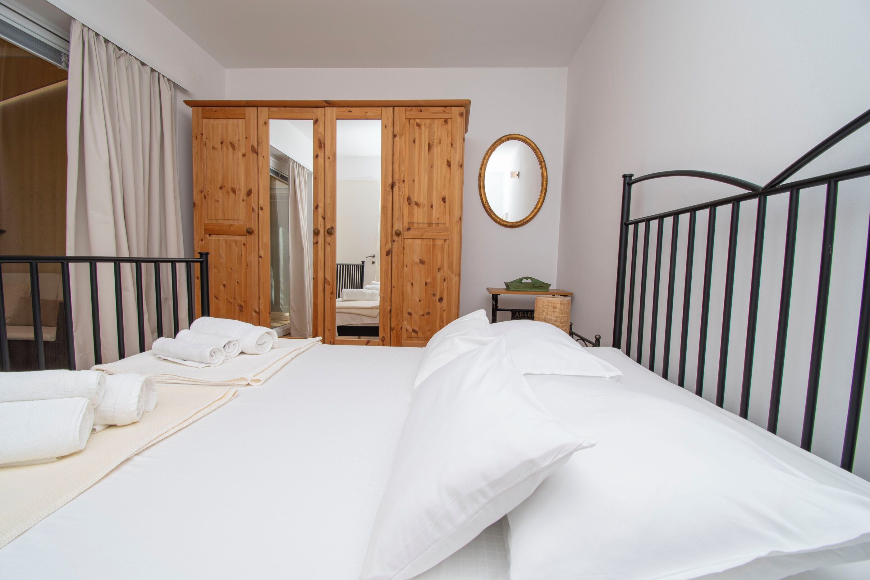 A bedroom with a bed, a wooden wardrobe, a mirror, and a bedside table with a lamp.