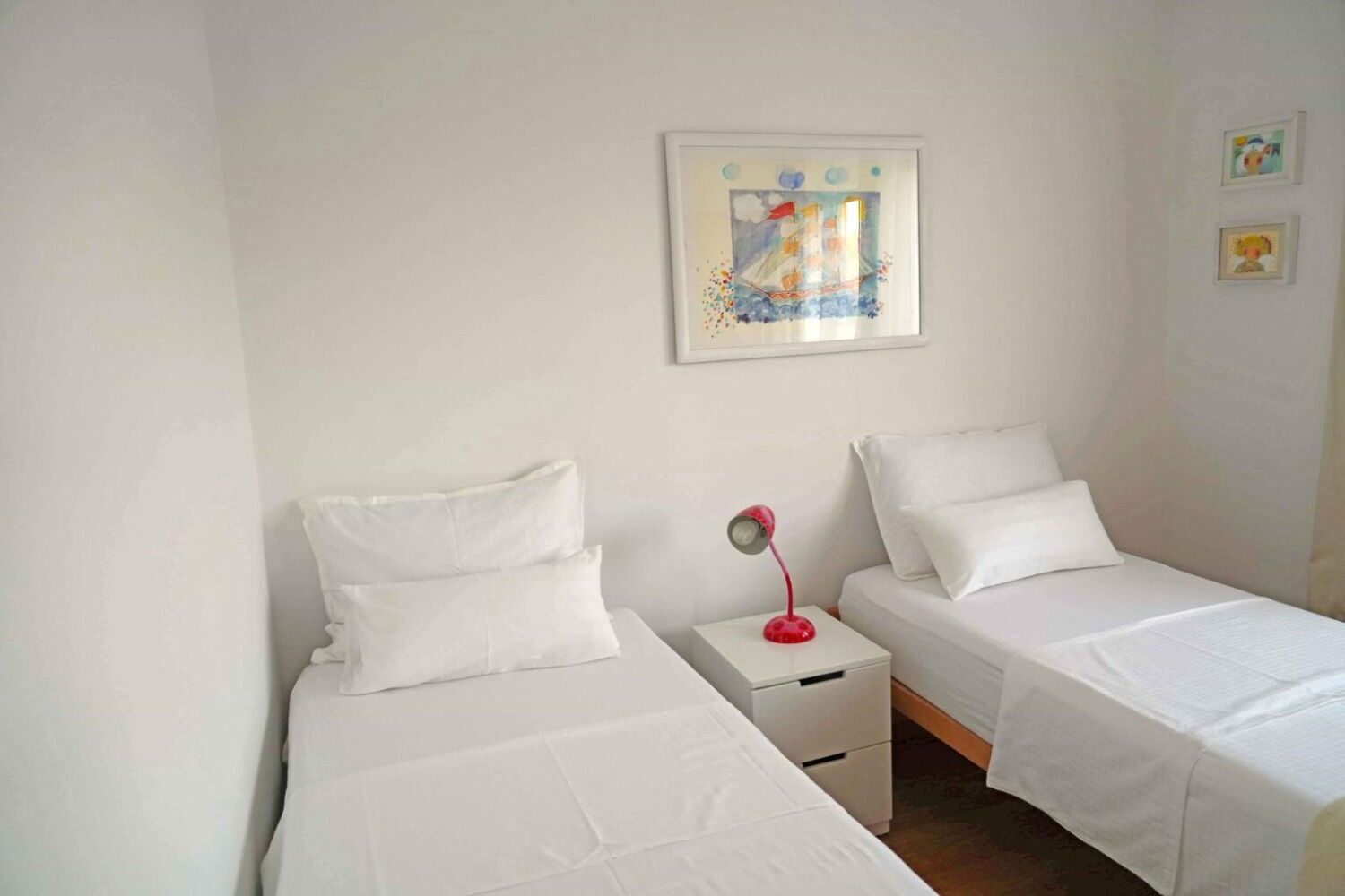 A room with two single beds, white bedding, a small bedside table with a red lamp, and a framed picture on the wall.