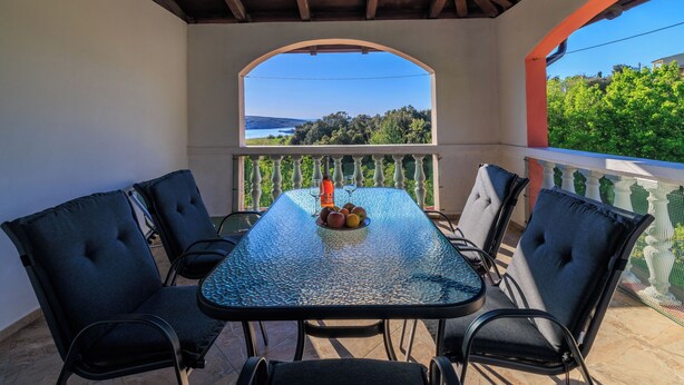 A covered outdoor patio with a table and chairs, a view of the landscape through an arched window, and a railing. - Apartments Grga (Lopar)