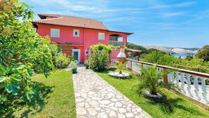 A pink villa with a stone pathway leading to the entrance, surrounded by greenery and a scenic view. - Apartments Grga (Lopar)