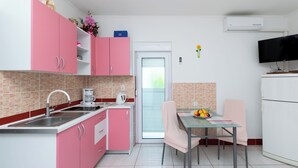 A modern kitchen with pink cabinets, a white countertop, and a dining area with a glass table and chairs. - Apartments Grga (Lopar)