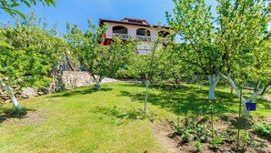 A house with a red roof and white walls, surrounded by a garden with trees and a stone wall. - Apartments Grga (Lopar)