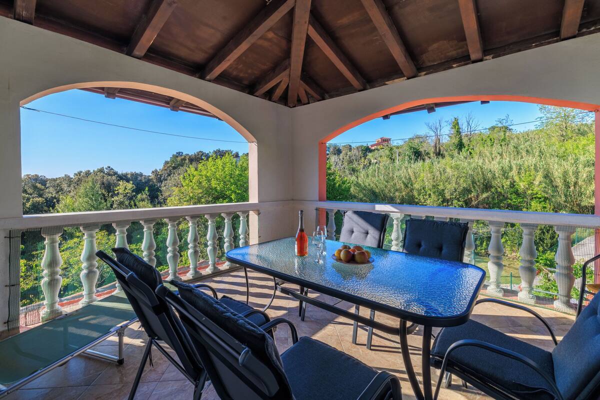 A covered outdoor seating area with a glass table, chairs, and a view of a hillside.