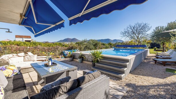 An outdoor pool area with lounge chairs, a covered seating area, and a view of mountains. - Apartments Holiday Home Book (Opuzen)