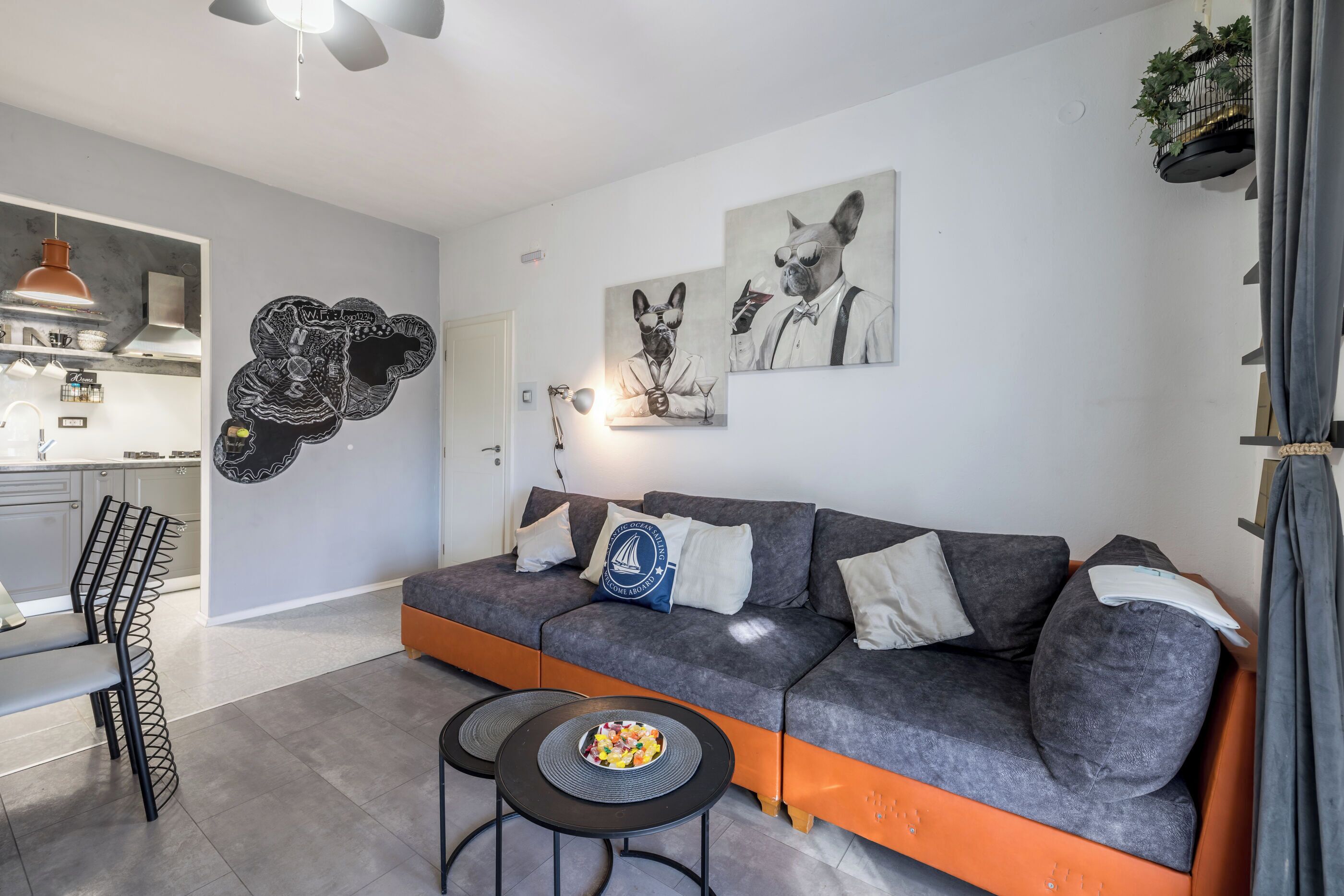Un salon moderne avec un canapé gris et orange, deux tables d’appoint noires et un saladier rempli de fruits. On y trouve également des œuvres d’art abstraites accrochées au mur et une plante suspendue.