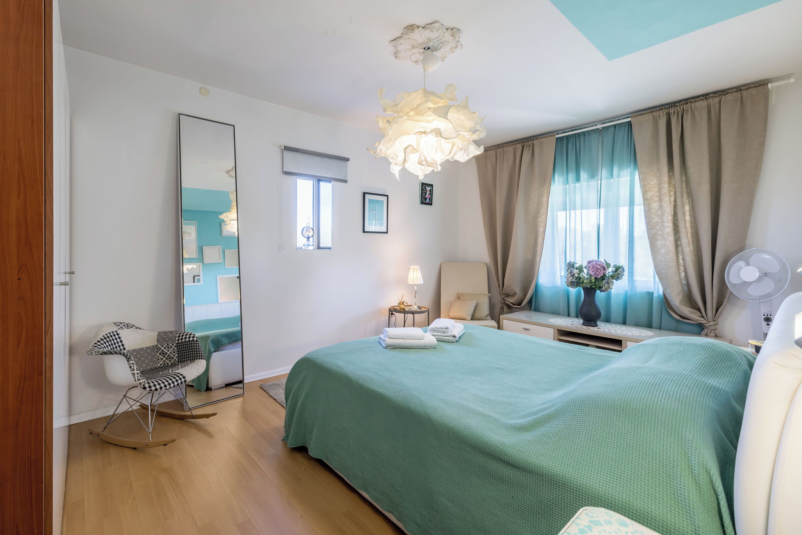 A bedroom with a green bedspread, a rocking chair, a bedside table with a lamp, and a large mirror.