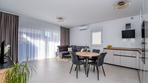 Een moderne woonkamer met een bank, een eettafel met stoelen, een aan de muur bevestigde televisie en een groot raam met gordijnen.