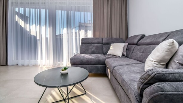 Een moderne woonkamer met een grijze modulaire bank, een ronde glazen salontafel en lichte gordijnen die natuurlijk licht doorlaten.
