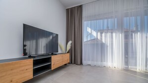 Een moderne woonkamer met een flatscreen televisie op een houten tv-meubel, grote ramen met lichte gordijnen en uitzicht op gebouwen.
