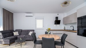 Een moderne woonkamer met een hoekbank, een ronde houten eettafel en zwarte stoelen. Er is een keuken met witte kasten en een zwarte oven.