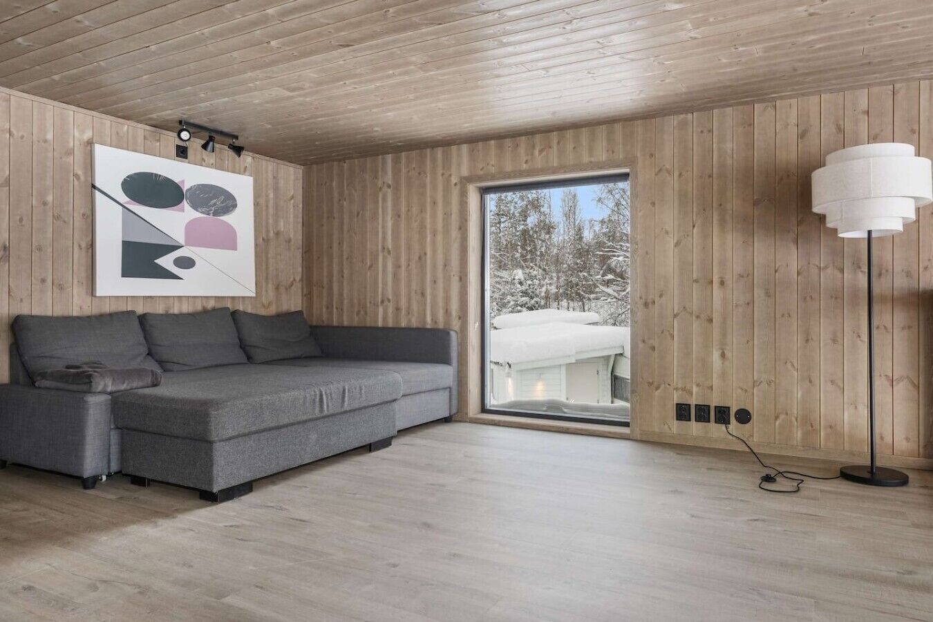A modern interior with a grey sofa, wooden walls, a large window, and a floor lamp.