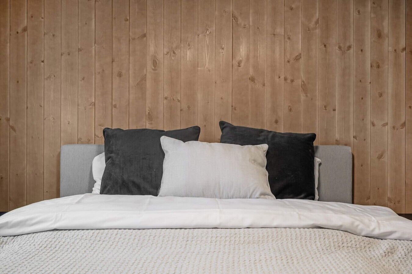 A bed with a grey headboard and white bedding, set against a wooden paneled wall.