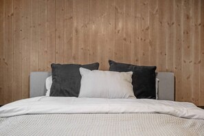 Cama con cabecera gris y ropa de cama blanca, frente a una pared con paneles de madera.