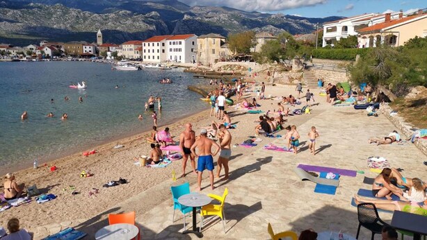 A beach with people sunbathing and swimming, a few tables and chairs, and buildings in the background. - House Anamaria (Posedarje)