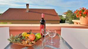 A table with a bottle of wine, two empty glasses, and a fruit platter on a balcony with a tiled roof and a view of a building in the background. - Apartments Lea Patrik Mara (Nin)