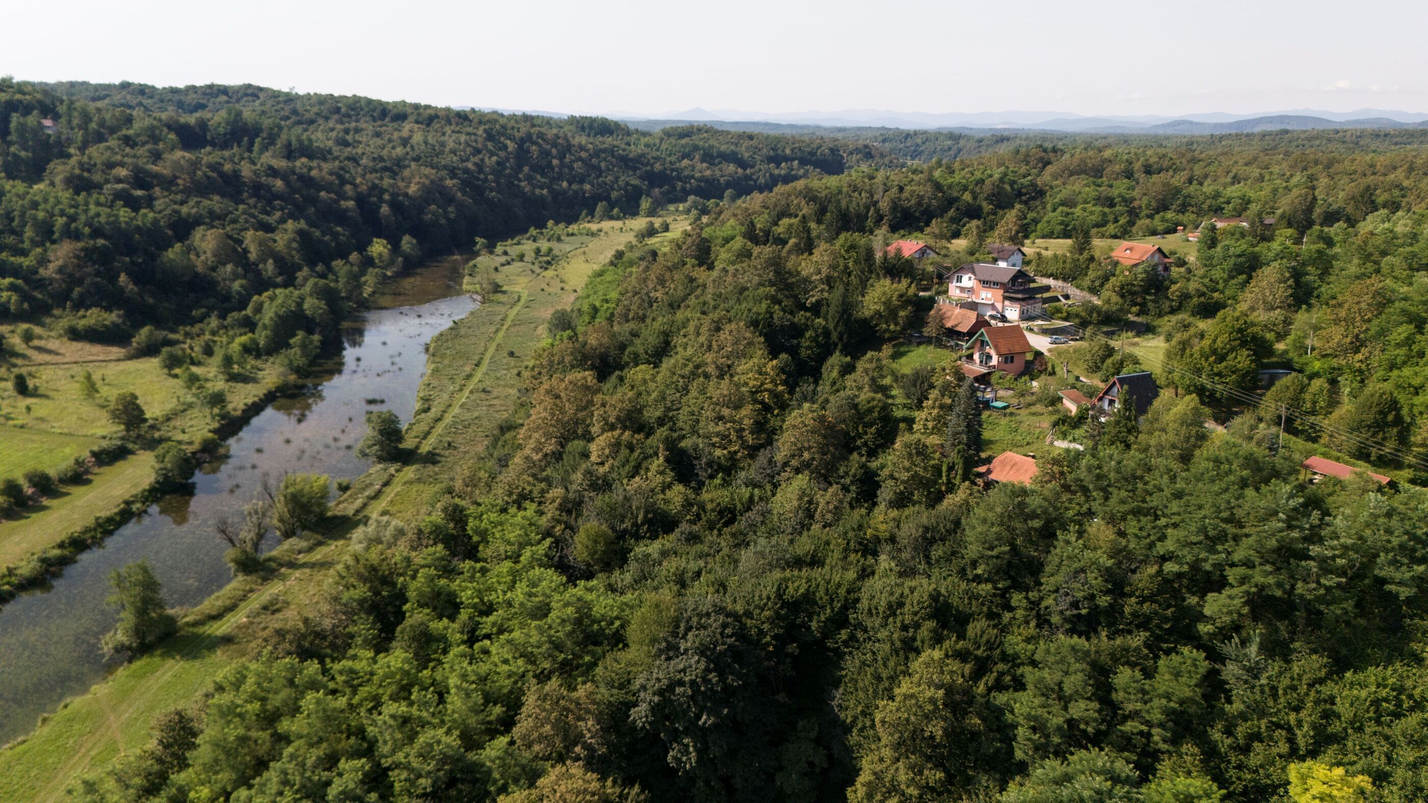 Un paysage rural avec une rivière, une forêt dense et un groupe de maisons aux toits rouges.