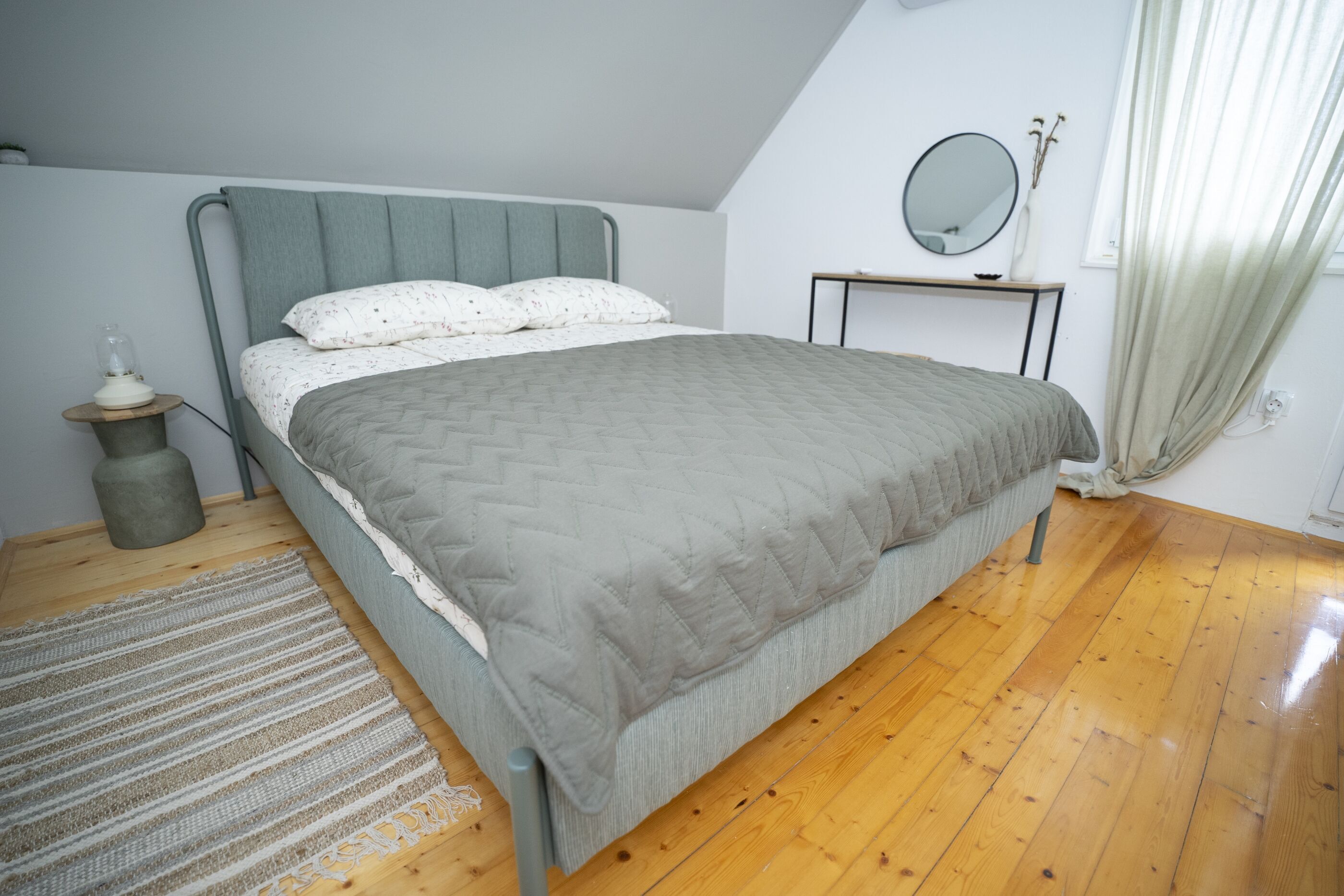 A bedroom with a bed, a bedside table, a mirror, and a window with curtains.