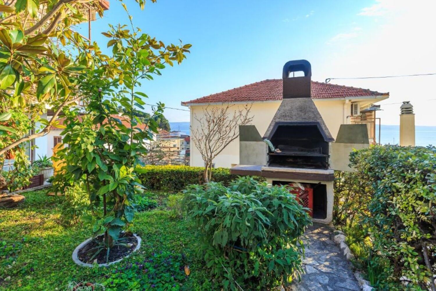 A house with a barbecue area, surrounded by greenery and a view of the sea.