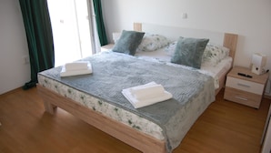 A neatly made bed with a floral-patterned bedspread and two pillows, a wooden nightstand with a lamp, and a wooden floor. - House Sun (Kastela)