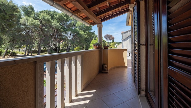A balcony with a tiled floor, wooden railing, and a view of trees and a building in the background. - Apartment Nikica (Supetar)