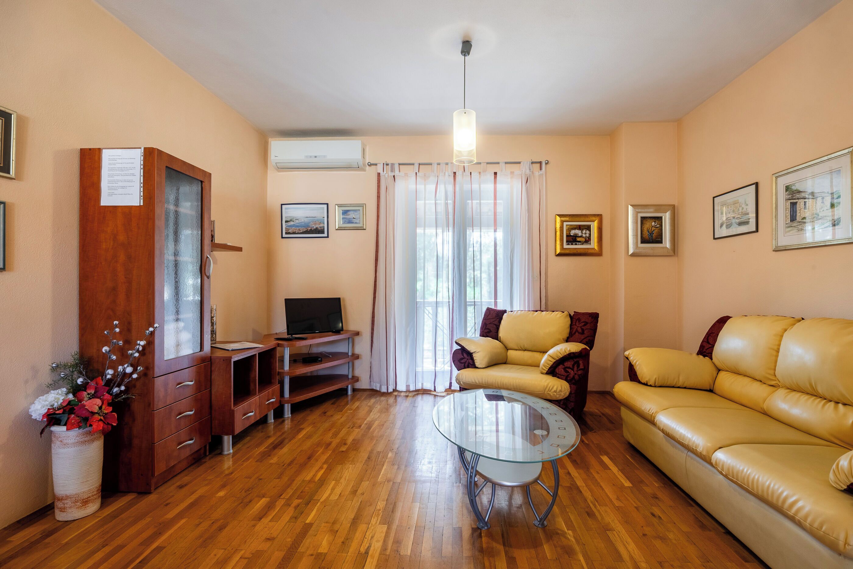 A living room with a wooden floor, a glass coffee table, a yellow sofa, a TV on a wooden stand, and a cabinet with a vase of flowers.