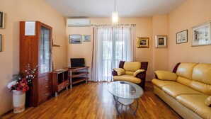 A living room with a wooden floor, a glass coffee table, a yellow sofa, a TV on a wooden stand, and a cabinet with a vase of flowers. - Apartment Nikica (Supetar)