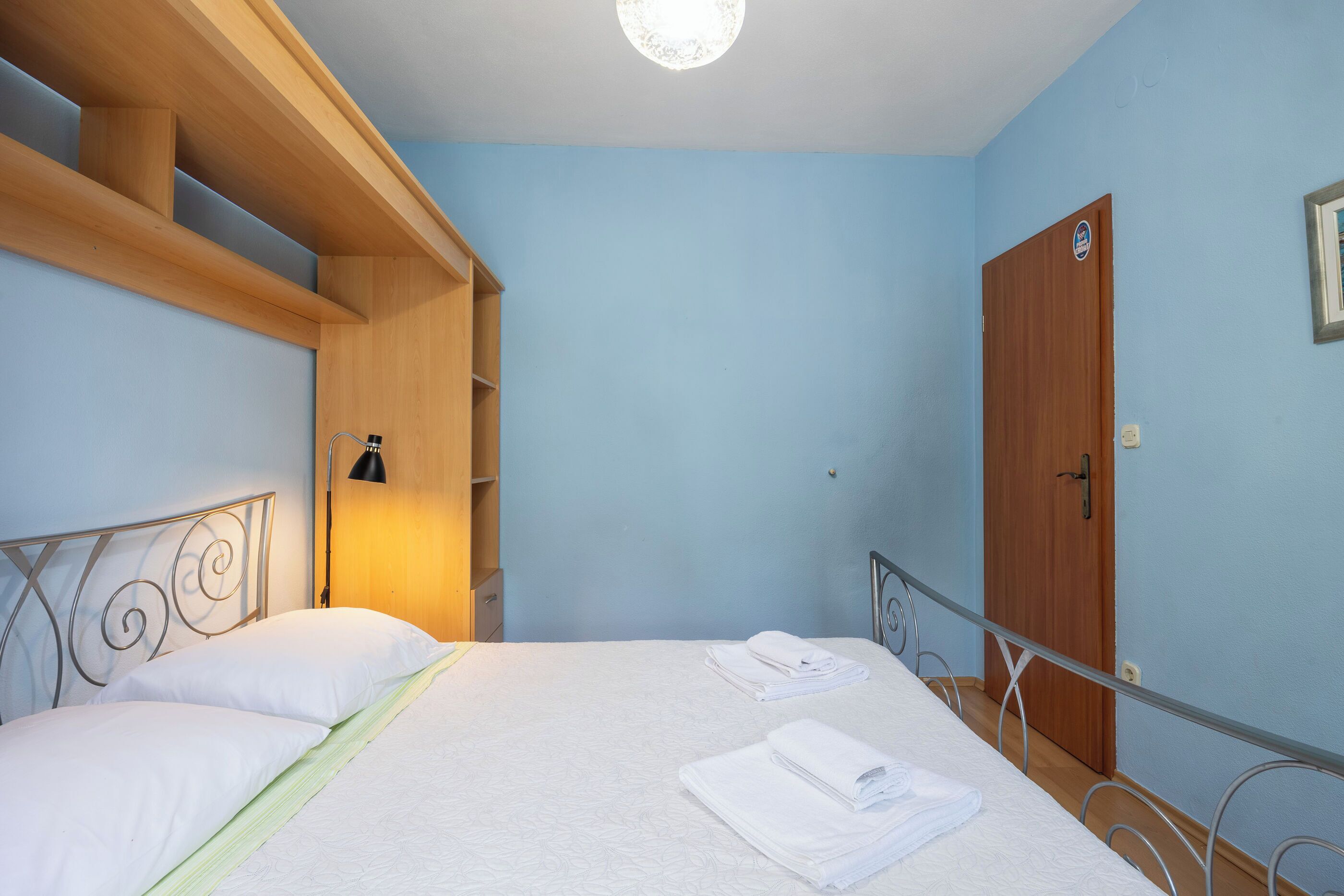 A bedroom with a bed, wooden headboard, a nightstand, a wall-mounted lamp, and a door.