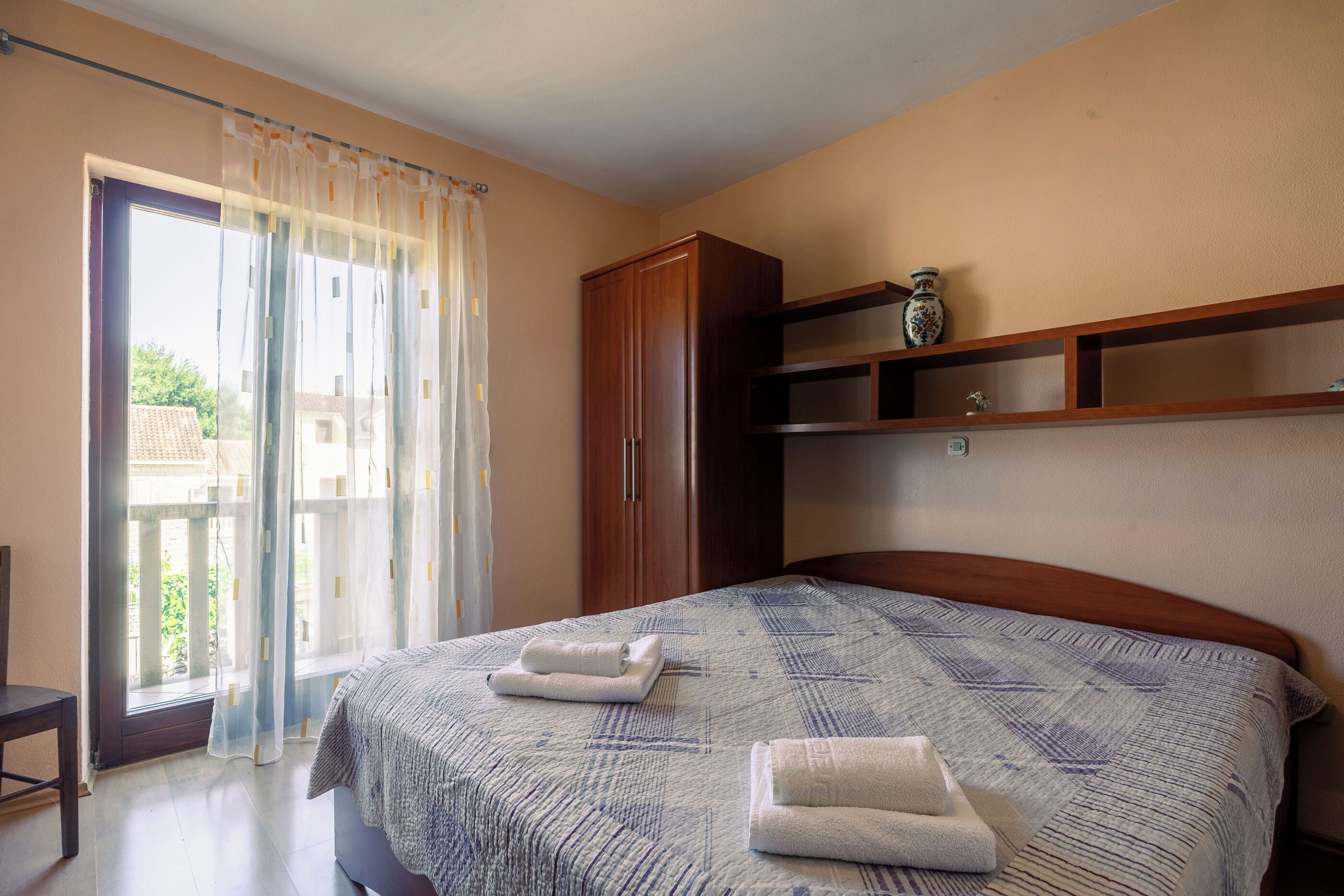 Una camera da letto con un letto, un armadio in legno, una finestra con tende e un piccolo ripiano con un vaso.