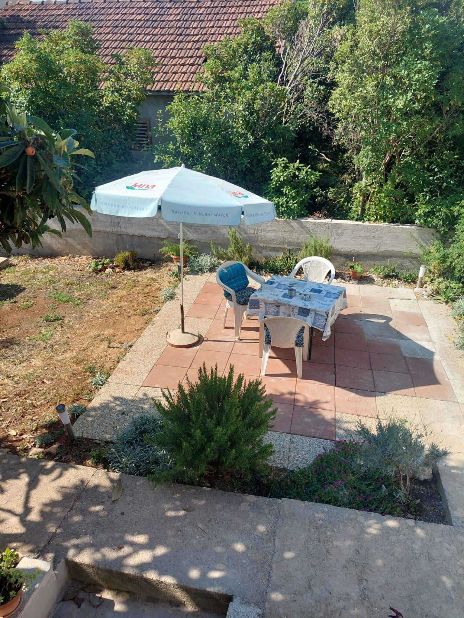 Eine Terrasse im Freien mit Tisch und Stühlen, einem schattenspendenden Sonnenschirm und einem Gartenbereich mit Pflanzen und einem kleinen Baum.