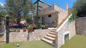 A stone house with a staircase leading to a covered porch, surrounded by potted plants and a grassy area. - Apartments Aura (Seget)