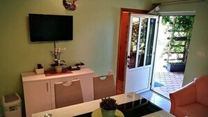 A dining area with a table, chairs, a TV mounted on the wall, and a door leading to an outdoor area with plants. - Apartments Aura (Seget)