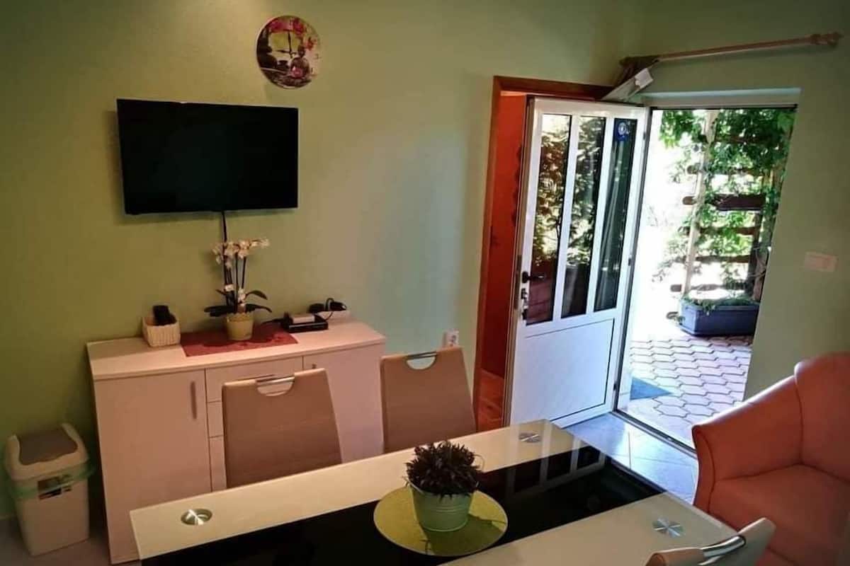 A dining area with a table, chairs, a TV mounted on the wall, and a door leading to an outdoor area with plants.