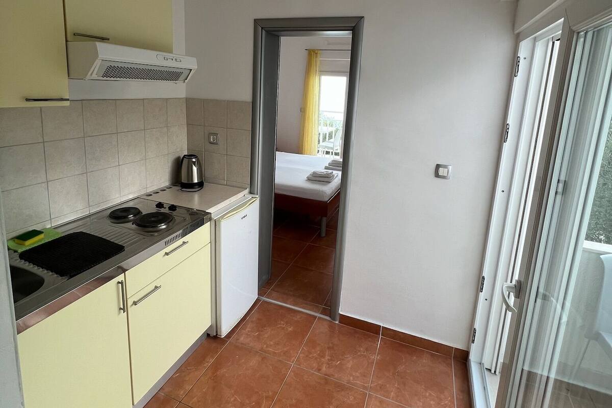 Cocina compacta con estufa, lavamanos y alacenas, que da paso a un dormitorio con cama y ventana.