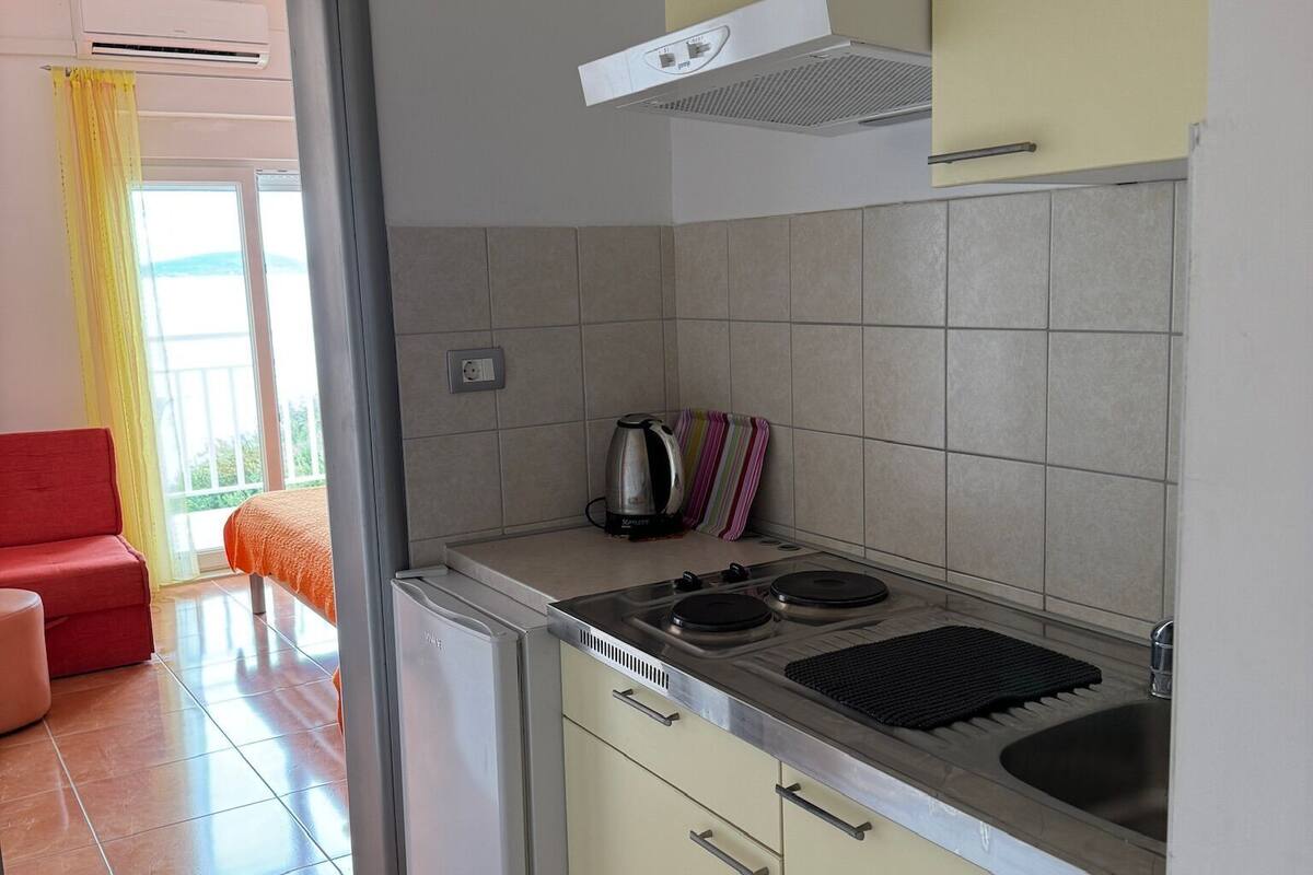 Una cocina compacta con refrigerador, estufa y pileta. En el fondo, se ve un pequeño comedor con una silla roja y una mesa.