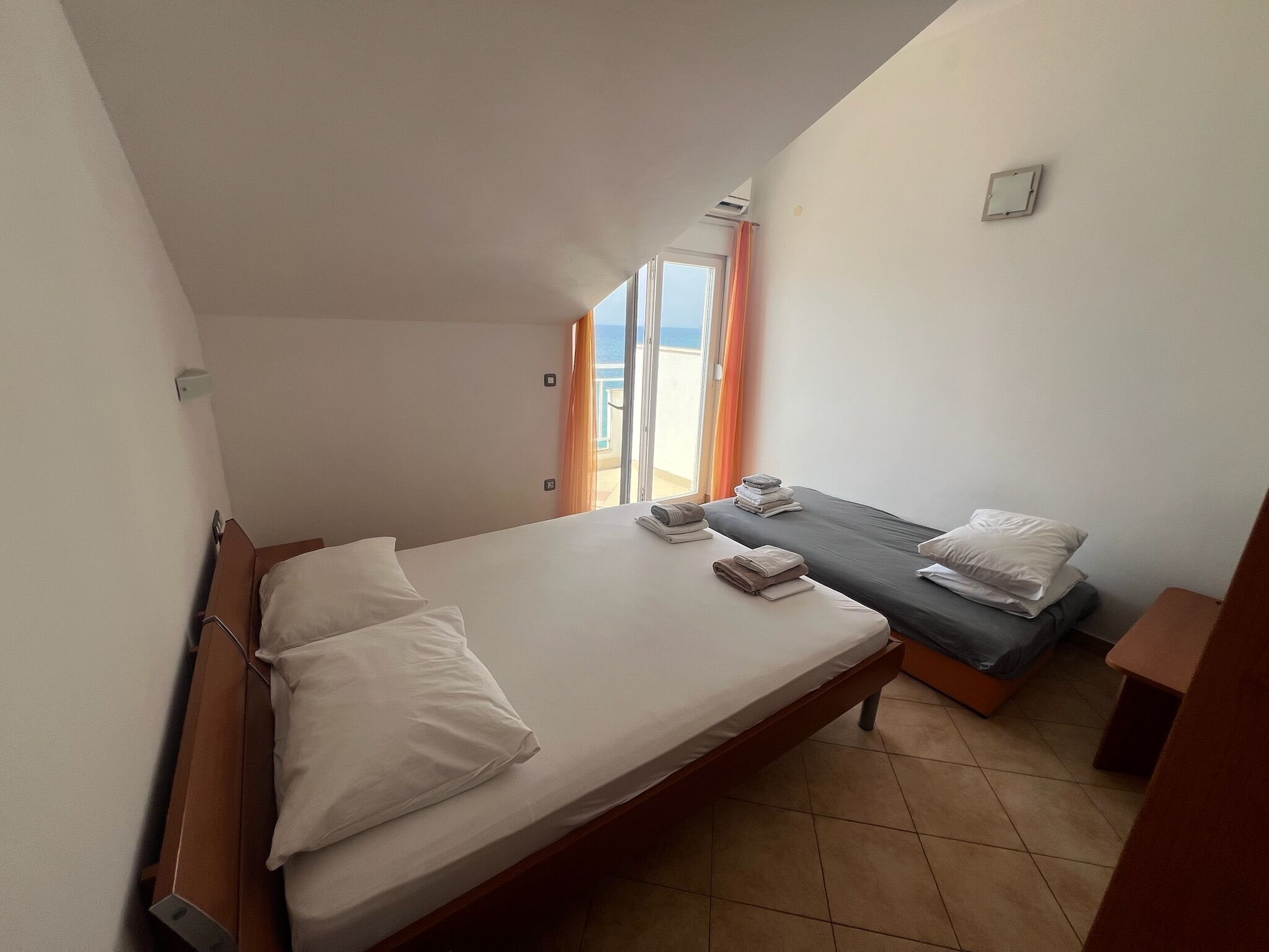A bedroom with a large bed, a smaller bed, and a desk. There is a window with curtains and a view of the outdoors.