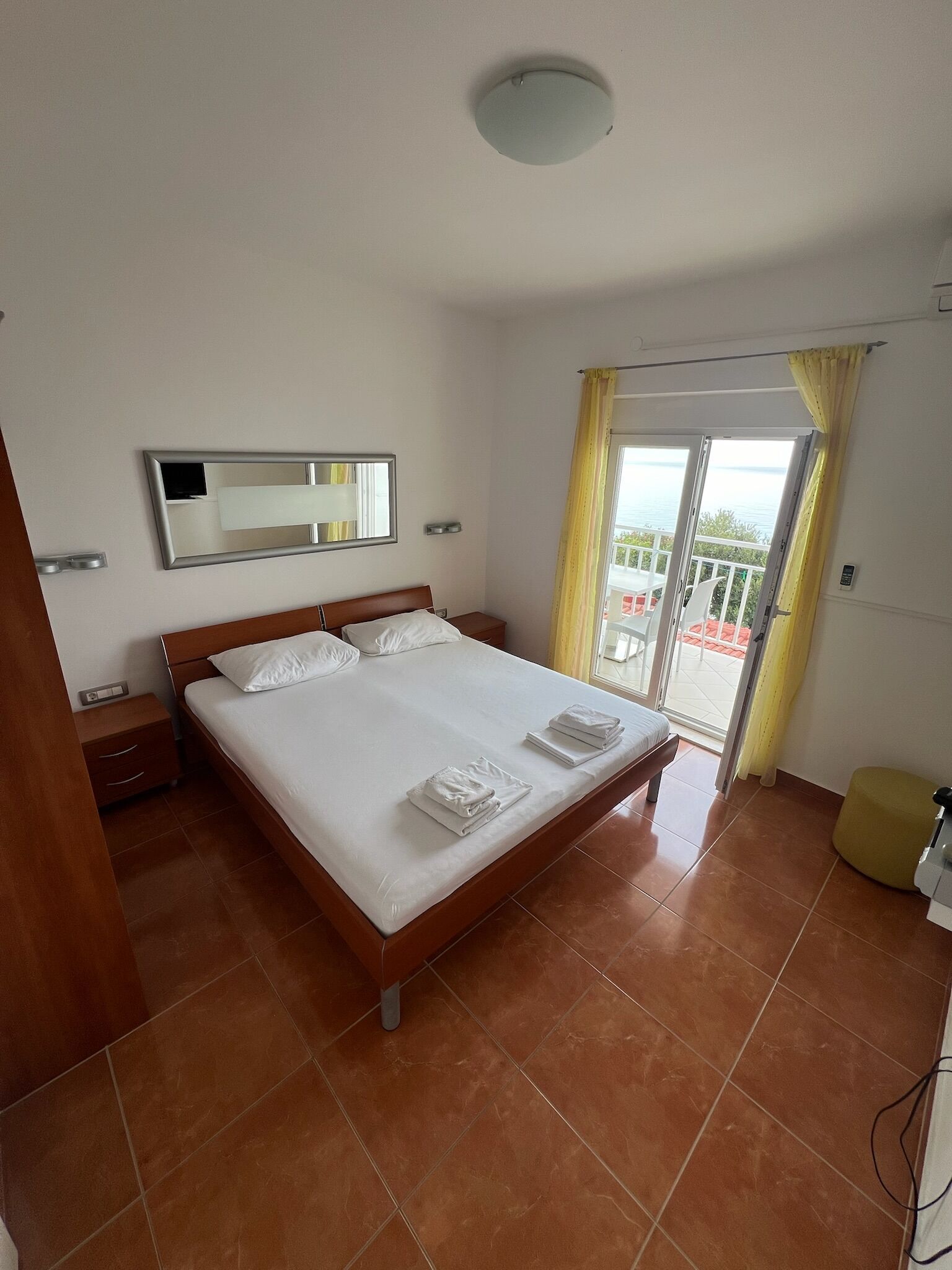 A hotel room with a bed, a nightstand, a mirror, and a view of the outside.