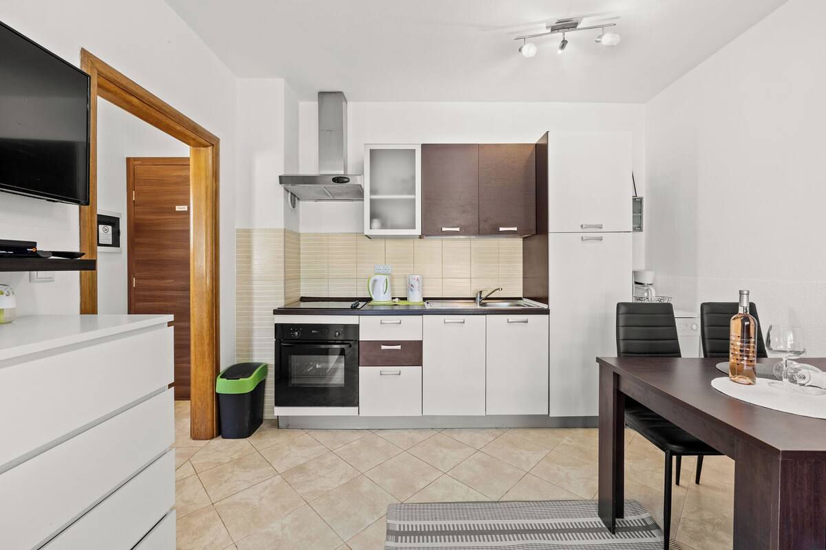 Een moderne keuken met witte kasten, een ingebouwde oven en een roestvrijstalen afzuigkap. Er staat een eettafel met stoelen en een kleine prullenbak.