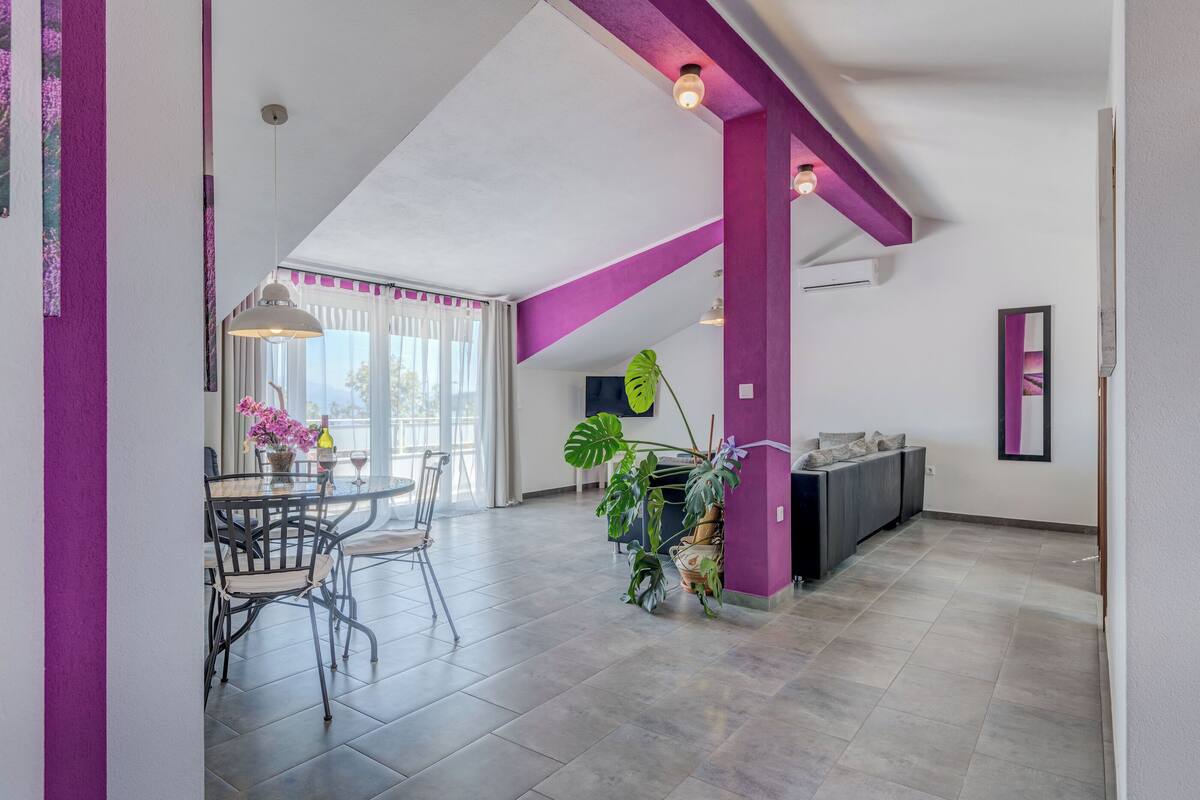 A spacious living area with a high ceiling, a dining table with chairs, and a large plant.