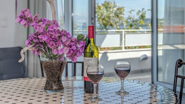 A table with a bottle of wine, two wine glasses, and a vase of purple flowers. - Apartments Deepwater (Split)