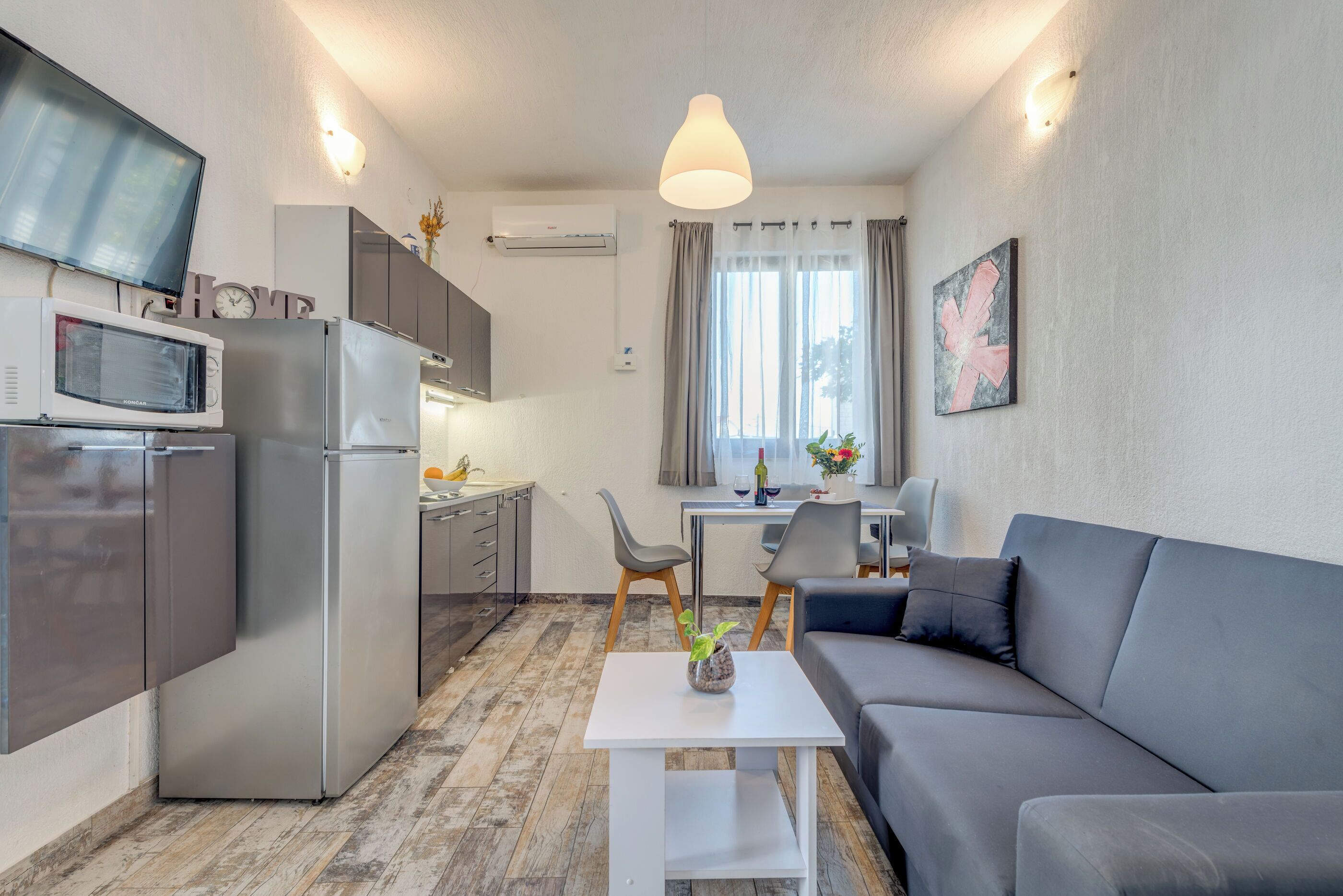 A compact living space with a kitchenette, a grey sofa, a white table, and a TV mounted on the wall.