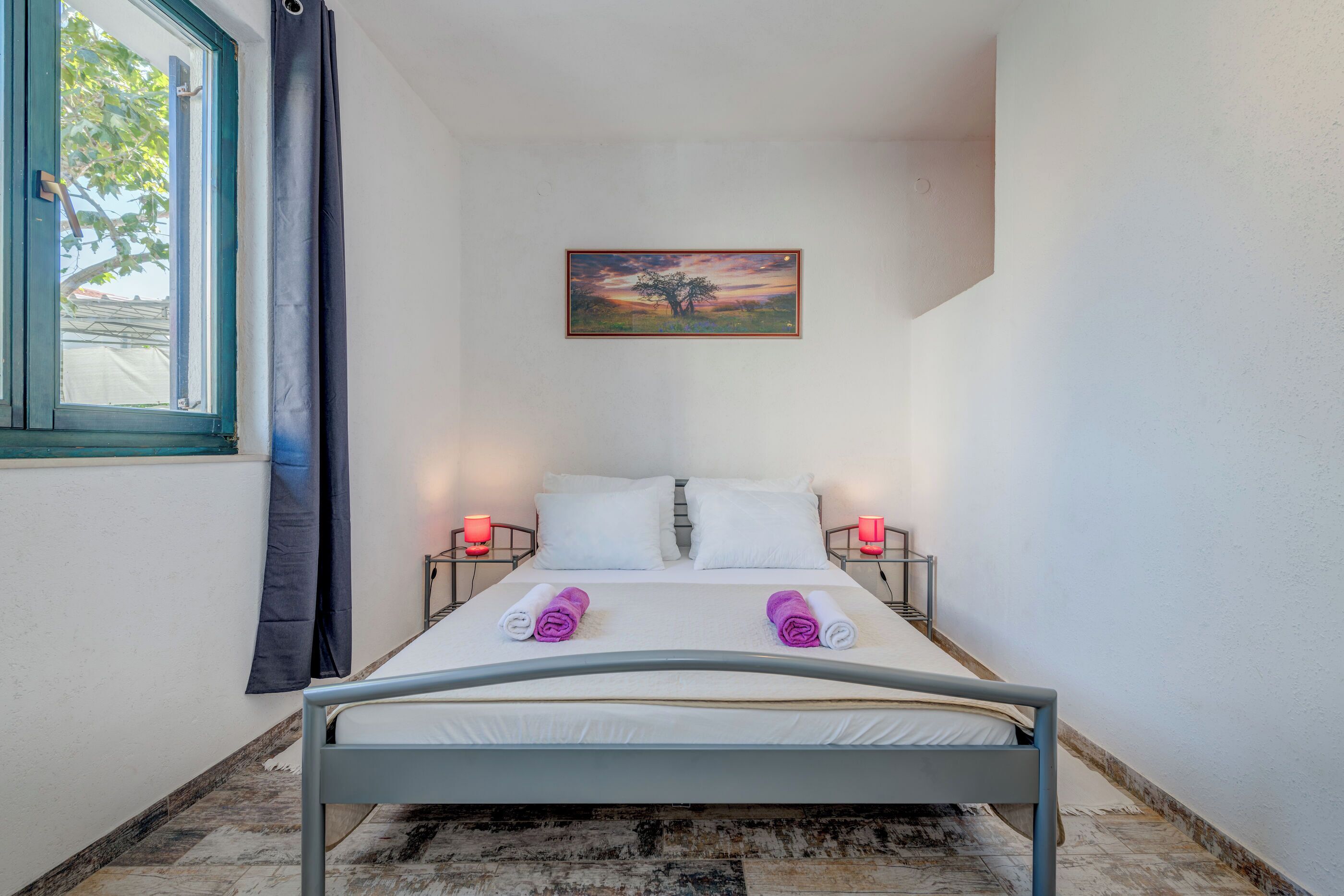 A neatly made bed with white linens and two rolled towels, a painting on the wall, and a window with curtains.