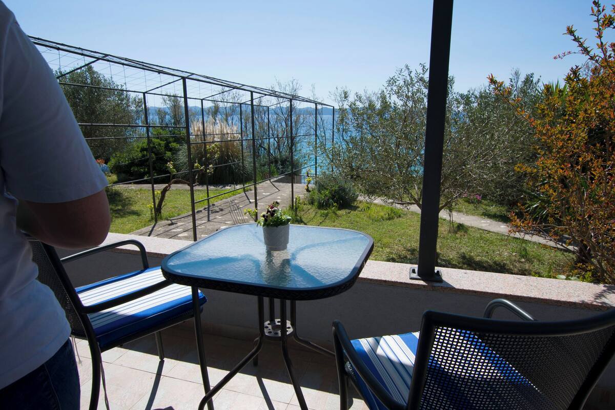 Un balcon avec une table et des chaises en verre, donnant sur un jardin et une étendue d’eau.