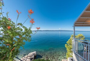 A coastal view with a clear blue sea, a rocky shoreline, and a building with a balcony overlooking the water. - Apartments Sun and Beach (Brela)