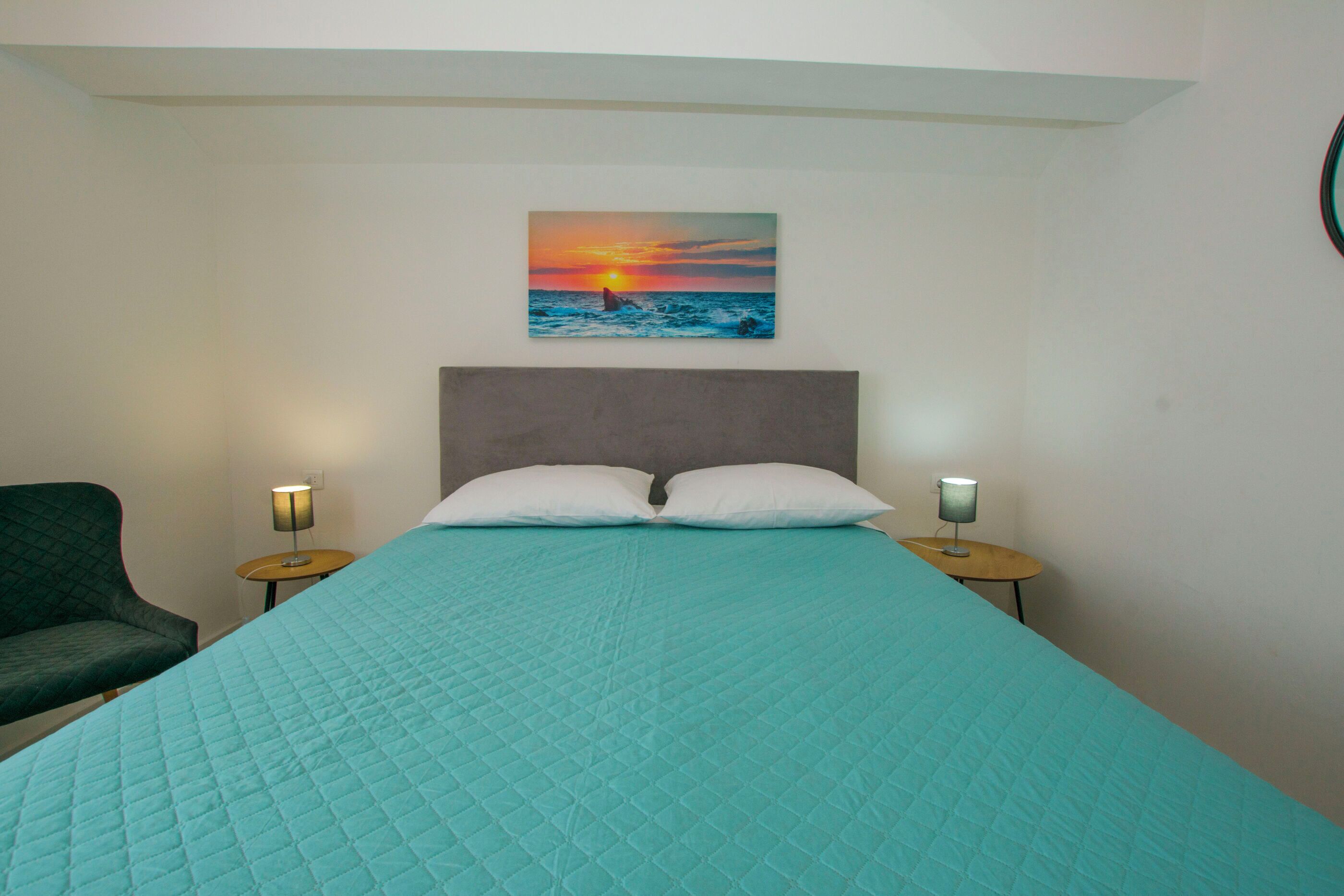 Un dormitorio con cama, dos lámparas de noche, una silla y un cuadro de una puesta de sol sobre el océano en la pared.