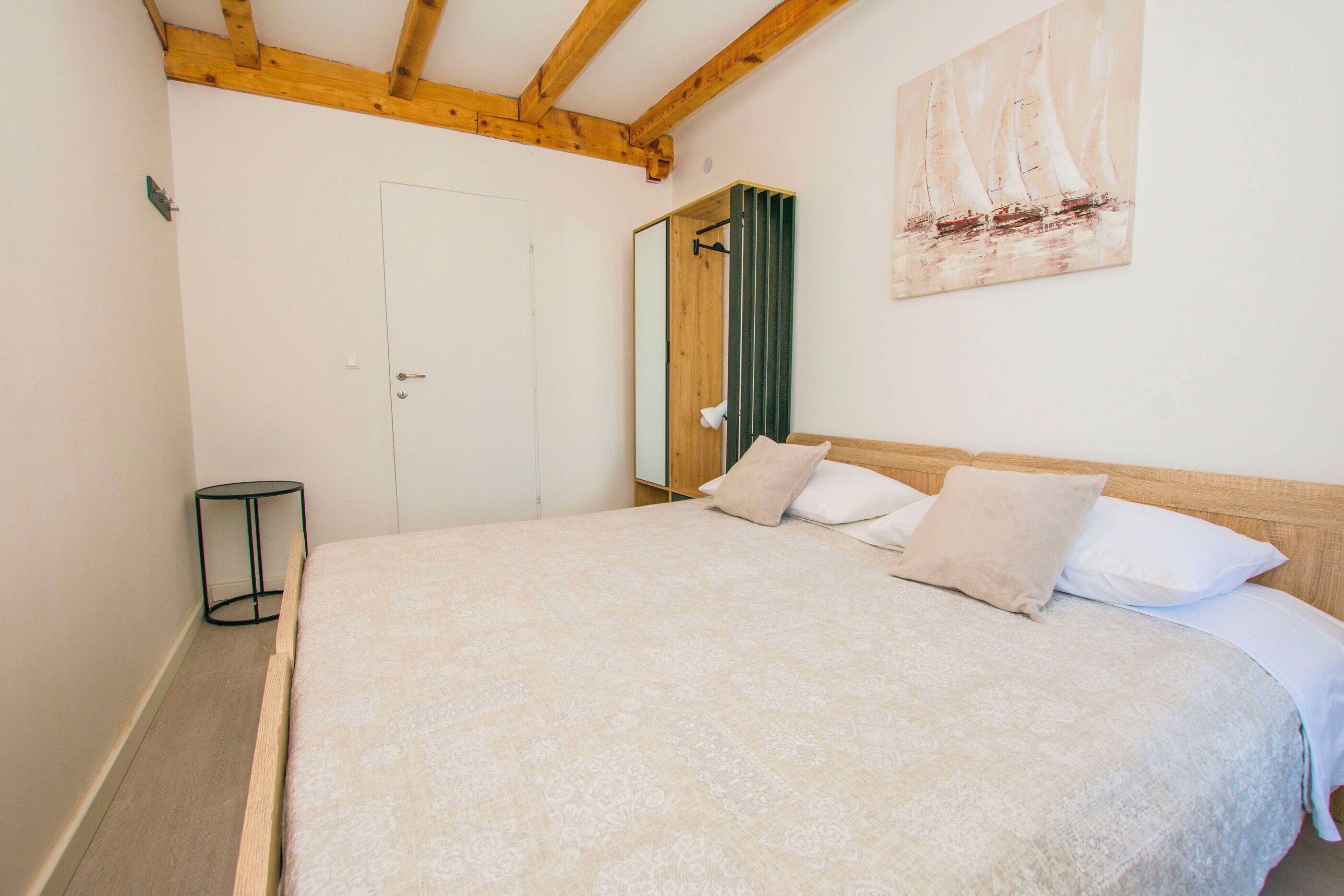A bedroom with a large bed, a wooden ceiling, a door, a mirror, and a painting of sailboats.